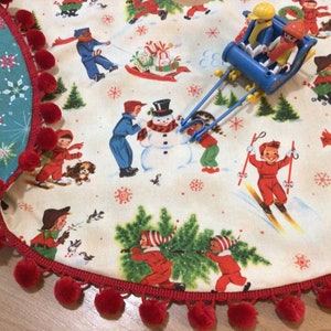 May include: A cream-colored Christmas tree skirt with a vintage winter scene featuring children playing in the snow. The skirt has a red pom-pom trim and a teal snowflake patterned border. A blue toy sled with figures is on top.