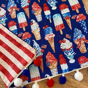 May include: A blue fabric featuring ice cream cones, ice lollies, and sundaes in red, white, and blue. A red and white striped fabric with pom-pom trim is on the left. The theme is patriotic.