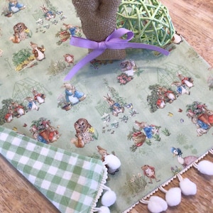 May include: A green and white checkered table runner with a Peter Rabbit print on one side. The runner has a white pom-pom trim and is perfect for a spring or Easter table setting.