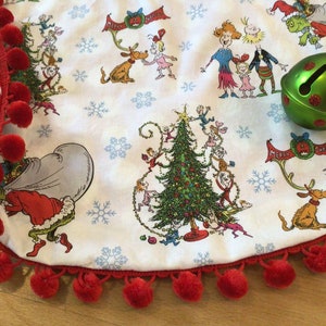 May include: A white Christmas tree skirt with a red and white polka dot trim and a red pom-pom border. The skirt features a print of characters from the book "How the Grinch Stole Christmas!" and a green bell with red polka dots.