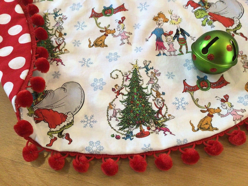 May include: A white Christmas tree skirt with a red and white polka dot trim and a red pom-pom border. The skirt features a print of characters from the book "How the Grinch Stole Christmas!" and a green bell with red polka dots.