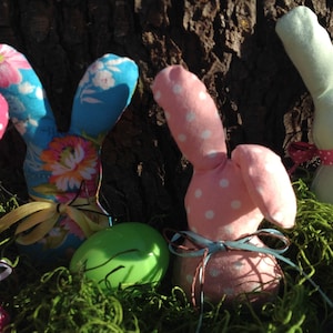 May include: Four handmade Easter bunny plush toys with different fabric patterns. The bunnies are pink with white polka dots, blue with floral print, green, and brown. The bunnies are sitting in a nest of green moss.
