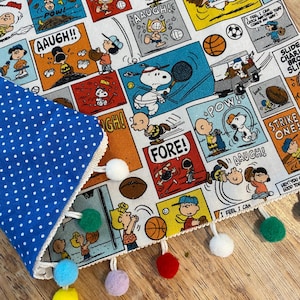 May include: A blue polka dot fabric with a white trim and colorful pom-poms. The fabric is paired with a comic strip featuring cartoon characters in various sports scenes, with text bubbles and action words.