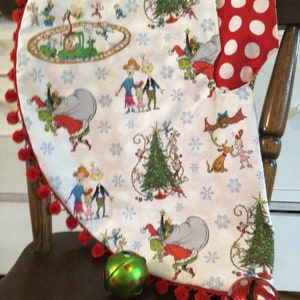 May include: A white Christmas tree skirt with a red pom-pom trim and a festive print of the Grinch and his friends. The skirt features a red and white polka dot fabric on the back.