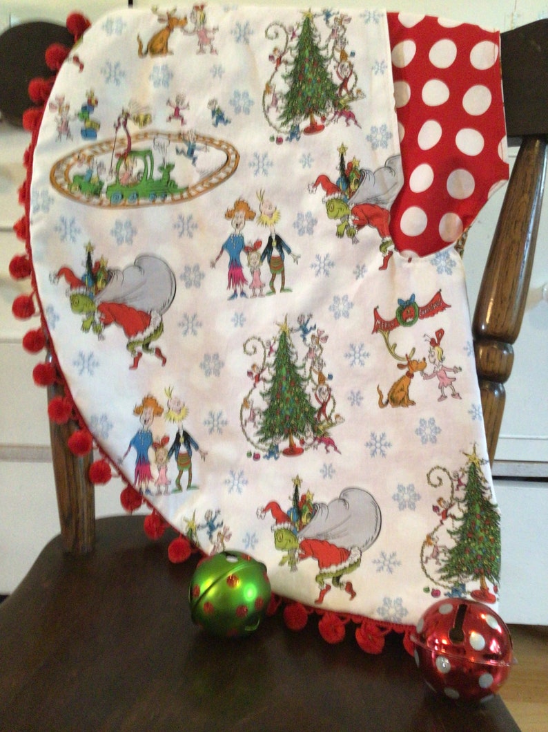 May include: A white Christmas tree skirt with a red pom-pom trim and a festive print of the Grinch and his friends. The skirt features a red and white polka dot fabric on the back.