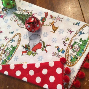 May include: A white table runner with a red and white polka dot border. The runner features a festive Christmas scene with the Grinch and his friends. The runner is trimmed with red pom-poms.