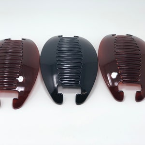 May include: Three plastic banana hair clips in shades of brown and black. Each clip has a curved, oval shape with a comb-like design and a clasp at the base. The clips are displayed against a white background.