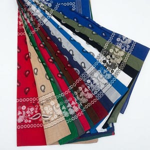 May include: A collection of square fabric bandanas arranged in a fan shape, featuring a variety of solid colors including red, tan, green, dark gray, maroon, blue, white, and black, all decorated with a traditional white paisley pattern.