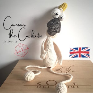 Op de afbeelding: Een gehaakt kaketoe speelgoed genaamd Caesar, met een gele kuif, een wit lichaam en een zwart gezicht. Het speelgoed zit op een houten oppervlak met een handgetekend zonnetje en de woorden "SO DEI JORI" eronder geschreven. De tekst "Caesar the Cockatoo pattern by Auntie Frankie" is zichtbaar in de linkerbovenhoek van de afbeelding.