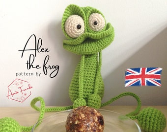 Puede incluir: Un juguete de rana de crochet verde llamado "Alex the frog" con grandes ojos blancos y una bola de energía marrón y blanca delante de él. El juguete está sentado en una superficie blanca con una pequeña bola verde de crochet a la izquierda y una bola verde de crochet más grande a la derecha. El texto "pattern by Auntie Franke" es visible en la imagen.