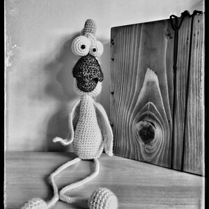 May include: A white crocheted toy with large eyes and a long beak, sitting on a wooden surface. The toy has long, thin legs and is wearing a small, crocheted hat.