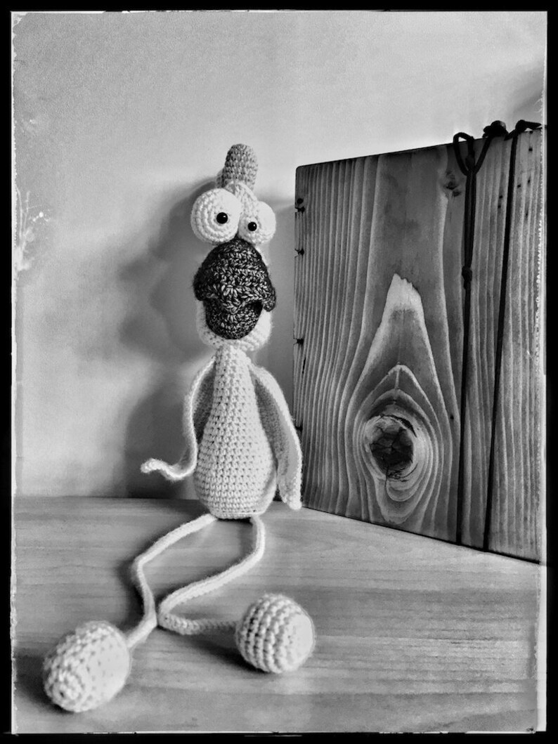 May include: A white crocheted toy with large eyes and a long beak, sitting on a wooden surface. The toy has long, thin legs and is wearing a small, crocheted hat.