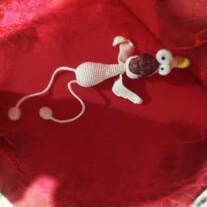 May include: White crocheted toy with a long tail, brown eyes, and a brown nose. The toy is lying on a red surface.