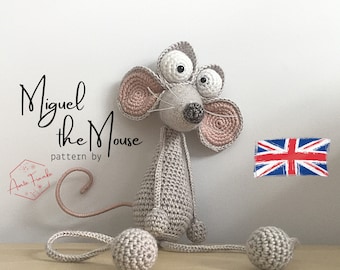 Puede incluir: Un juguete de ratón gris de crochet con grandes orejas rosas y grandes ojos blancos. El ratón está sentado sobre una superficie de madera con dos bolas grises. El texto "Miguel the Mouse pattern by Auntie Frankie" es visible en la imagen.