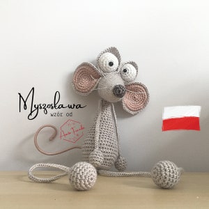 May include: A crocheted grey mouse with large pink ears and white eyes. The mouse has a long tail and is sitting on a wooden surface. The text "Myszosława wzór od Aunte Franko" is visible in the image.