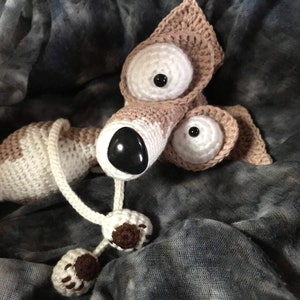 Puede incluir: Un perro de peluche tejido a crochet con grandes ojos blancos y una nariz marr&oacute;n. El perro est&aacute; acostado sobre una tela gris y blanca.