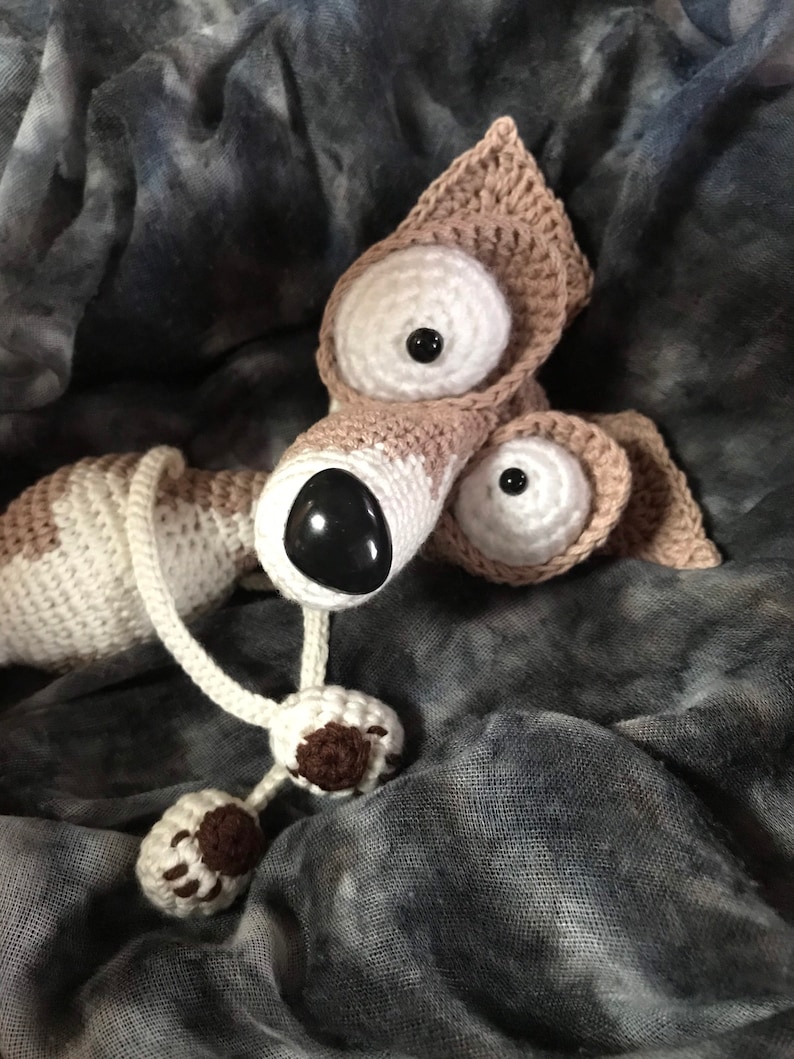 Puede incluir: Un perro de peluche tejido a crochet con grandes ojos blancos y una nariz marr&oacute;n. El perro est&aacute; acostado sobre una tela gris y blanca.