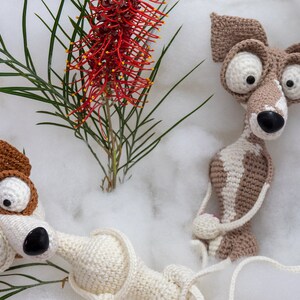 Puede incluir: Dos juguetes de perro de crochet con ojos grandes y narices marrones. Los perros est&aacute;n sentados sobre una superficie blanca y esponjosa con una flor roja en el fondo.