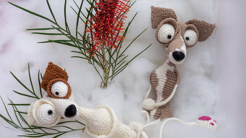 Puede incluir: Dos juguetes de perro de crochet con ojos grandes y narices marrones. Los perros est&aacute;n sentados sobre una superficie blanca y esponjosa con una flor roja en el fondo.