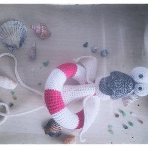 May include: A crocheted stuffed animal with a white and pink life preserver, gray head, and large white eyes. The toy is surrounded by seashells and small glass beads.
