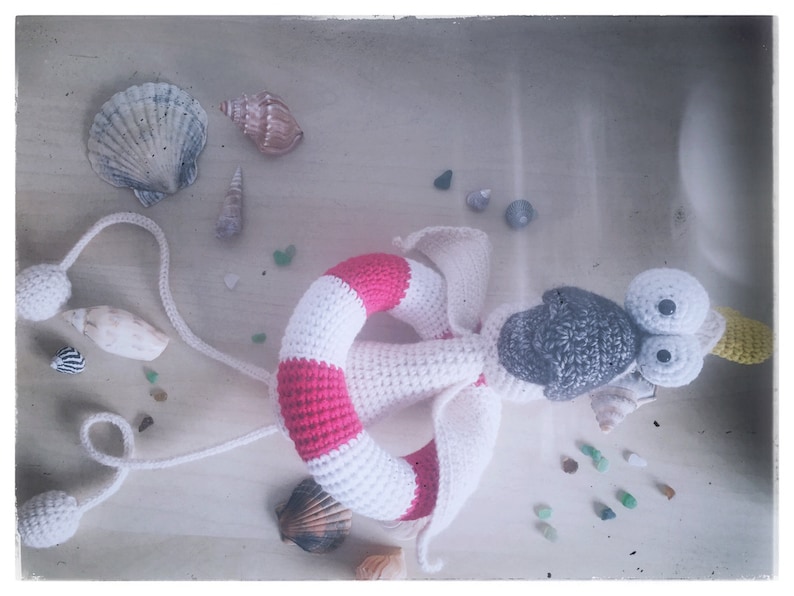 May include: A crocheted stuffed animal with a white and pink life preserver, gray head, and large white eyes. The toy is surrounded by seashells and small glass beads.