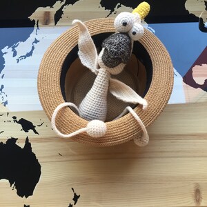 May include: A crocheted toy animal with large eyes and a yellow crown sits inside a straw hat. The hat is on a wooden surface with a black and white world map printed on it.