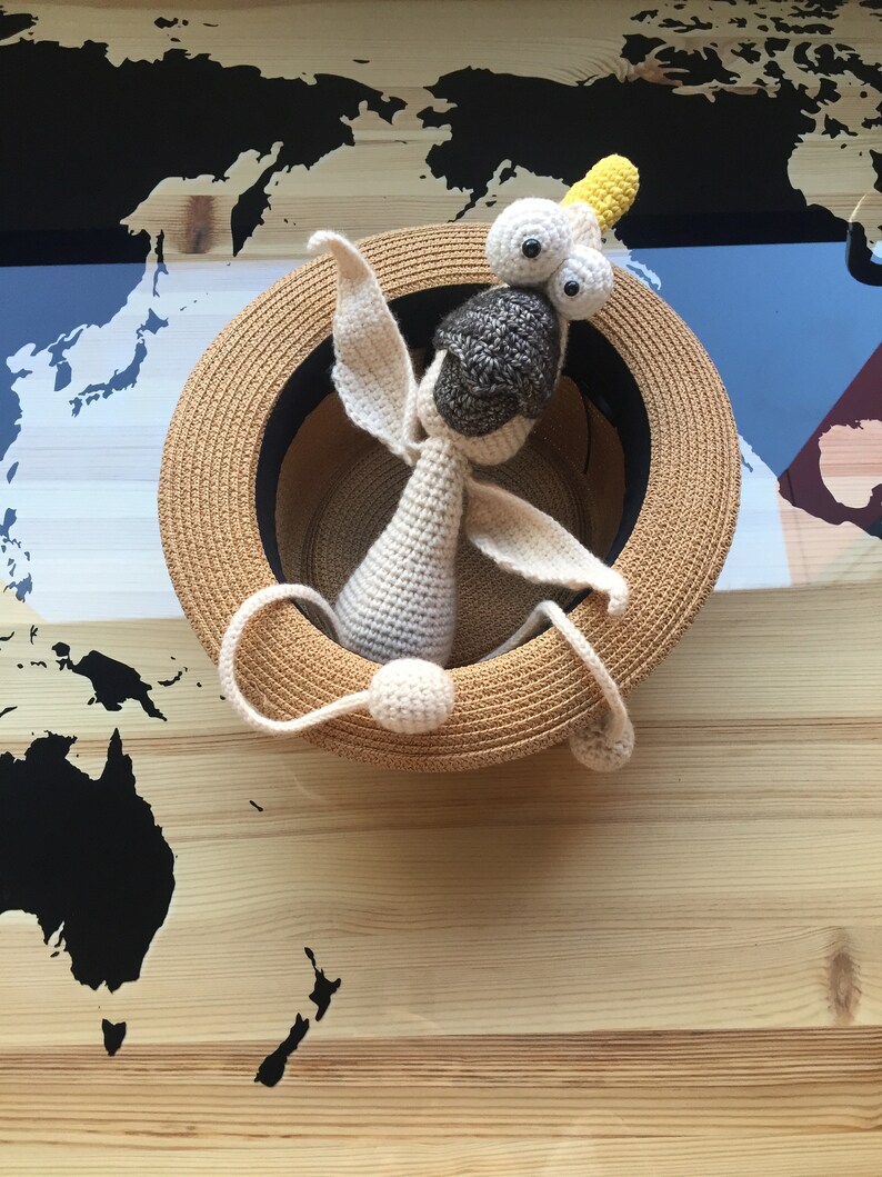 May include: A crocheted toy animal with large eyes and a yellow crown sits inside a straw hat. The hat is on a wooden surface with a black and white world map printed on it.