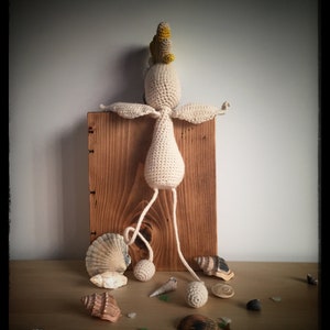 May include: A crocheted doll with a yellow and gray crown, standing on a wooden box. The doll has long legs and is surrounded by seashells.