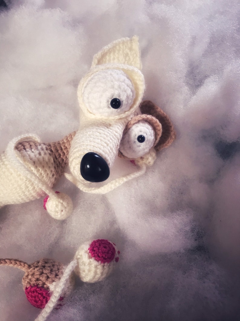 Puede incluir: Un juguete de perro de crochet blanco con detalles marrones y grandes ojos negros. El juguete tiene detalles rosas en las orejas y las patas.