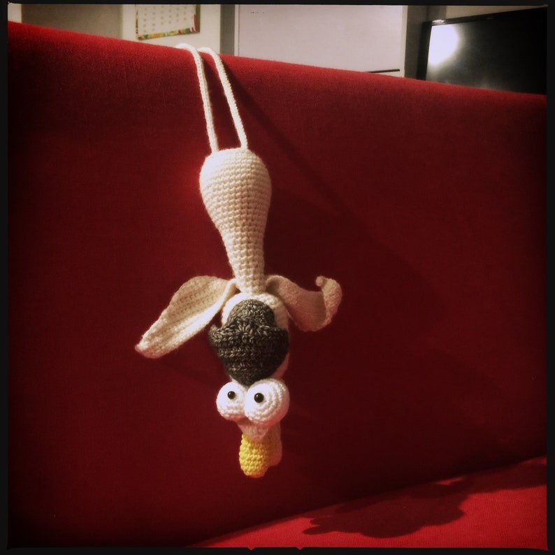 May include: A crocheted stuffed animal hanging upside down from a red couch. The animal has a white body, black eyes, and a brown hat. It has a yellow beak and white wings.