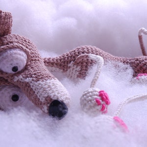 Puede incluir: Un juguete de perro de crochet hecho con hilo marr&oacute;n y blanco, acostado sobre una superficie blanca y esponjosa. El juguete tiene ojos grandes y una nariz rosa. El perro sostiene una pelota rosa y blanca en su pata.