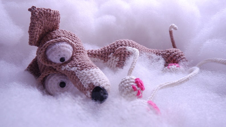 Puede incluir: Un juguete de perro de crochet hecho con hilo marr&oacute;n y blanco, acostado sobre una superficie blanca y esponjosa. El juguete tiene ojos grandes y una nariz rosa. El perro sostiene una pelota rosa y blanca en su pata.