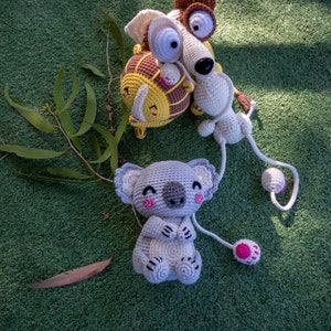 Puede incluir: Tres animales de peluche tejidos a crochet sobre una superficie verde. Un koala gris con detalles rosas, un perro marr&oacute;n y blanco y un le&oacute;n amarillo y marr&oacute;n.