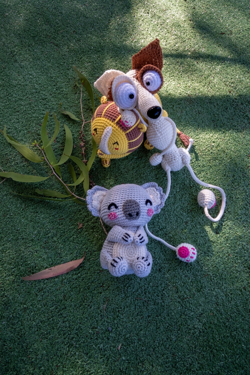 Puede incluir: Tres animales de peluche tejidos a crochet sobre una superficie verde. Un koala gris con detalles rosas, un perro marr&oacute;n y blanco y un le&oacute;n amarillo y marr&oacute;n.