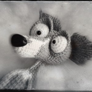 Puede incluir: Un animal de peluche de crochet blanco y negro con ojos grandes y un hocico largo. El animal est&aacute; acostado sobre una superficie blanca.
