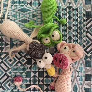 May include: Three crocheted stuffed animals: a green alien with large eyes, a brown and white dog with large eyes, and a pink pig with large eyes. The animals are on a blue and white patterned fabric.