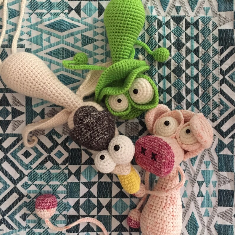 May include: Three crocheted stuffed animals: a green alien with large eyes, a brown and white dog with large eyes, and a pink pig with large eyes. The animals are on a blue and white patterned fabric.