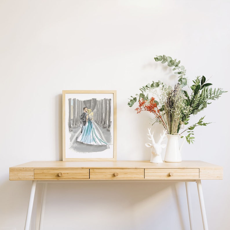 K&ouml;nnte beinhalten: Eine gerahmte Illustration eines Paares in einem Hochzeitskleid und Anzug, das in einer Waldlandschaft steht. Die Illustration ist in Schwarz, Wei&szlig; und Blau gehalten und wird auf einem Holztisch mit einer wei&szlig;en Vase und einer wei&szlig;en Hirschfigur ausgestellt.