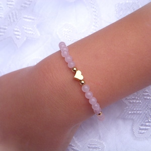 Peut inclure: Un bracelet en perles roses avec un pendentif en forme de cœur doré. Le bracelet est porté au poignet.