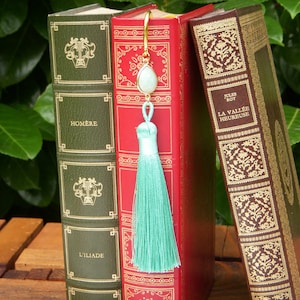 May include: A turquoise tassel bookmark with a gold metal chain and a light green gemstone. The bookmark is hanging from a stack of three hardback books. The books have gold lettering on their spines. The book on the left has the title "Homère" and "L'Iliade". The book in the middle has a red cover. The book on the right has the title "Jules Roy" and "La Vallée Heureuse".