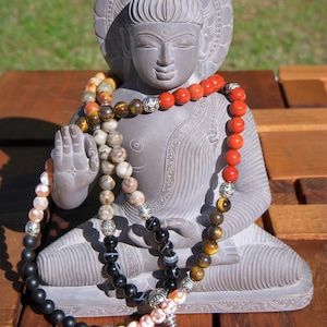 Peut inclure: Une statue de Bouddha en pierre grise avec un collier de perles en pierres noires, rouges, brunes et blanches. Le collier a un charme argenté à l'extrémité.