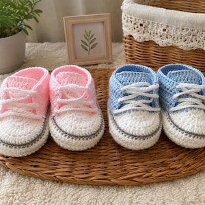 Puede incluir: Dos pares de patucos de bebé tejidos a mano con diseño de zapatillas. Un par es rosa claro y blanco, y el otro es azul claro y blanco. Cada zapato tiene cordones blancos, una puntera blanca y una fina raya gris a lo largo de la suela.