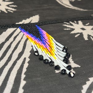 May include: A beaded choker necklace featuring a black band and a central fringe pendant. The fringe is composed of rows of small beads in shades of blue, pink, orange, yellow, and white, ending in clear white tubes tipped with small black beads.