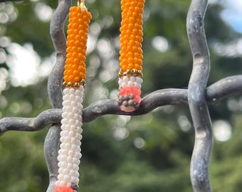 May include: A pair of dangling earrings featuring a cylindrical beaded design. The beads are arranged in segments of bright orange, white, and a small section of pink and grey at the base. Each earring is attached to a gold-toned metal lever back hook.