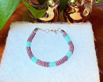 May include: A beaded bracelet with alternating sections of purple and turquoise beads. The bracelet is displayed on a white surface, with two gold bird figurines in the background. The bracelet has a silver clasp.