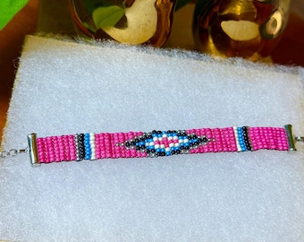 May include: A beaded bracelet with a vibrant pink background and a central geometric design in black, white, and blue. The bracelet has silver-toned metal end caps and a delicate chain clasp. The bracelet is displayed on a white surface.