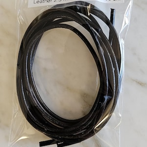 Könnte beinhalten: Schwarze, runde Lederschnur, 2,5 mm dick und 90 cm lang, verpackt in einem transparenten Plastikbeutel. Der Text auf dem Beutel lautet "Leather 2.5 mm round Black 90 cm".