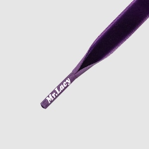 Puede incluir: Primer plano de una puntera de cordón de terciopelo morado. La puntera del cordón tiene el texto "Mr. Lacy" impreso en blanco. El cordón es de color morado intenso y parece estar hecho de un material suave y aterciopelado.