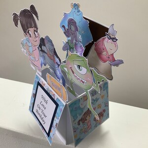 May include: A pop-up birthday card with a blue and white patterned background. The card features characters from the movie Monsters Inc., including Mike Wazowski, Sulley, Boo, and Randall. The card is open and the characters are popping out. The card also has a message that reads 'With Love On Your Birthday.'