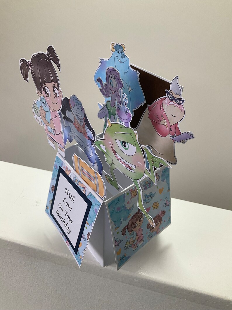 May include: A pop-up birthday card with a blue and white patterned background. The card features characters from the movie Monsters Inc., including Mike Wazowski, Sulley, Boo, and Randall. The card is open and the characters are popping out. The card also has a message that reads 'With Love On Your Birthday.'